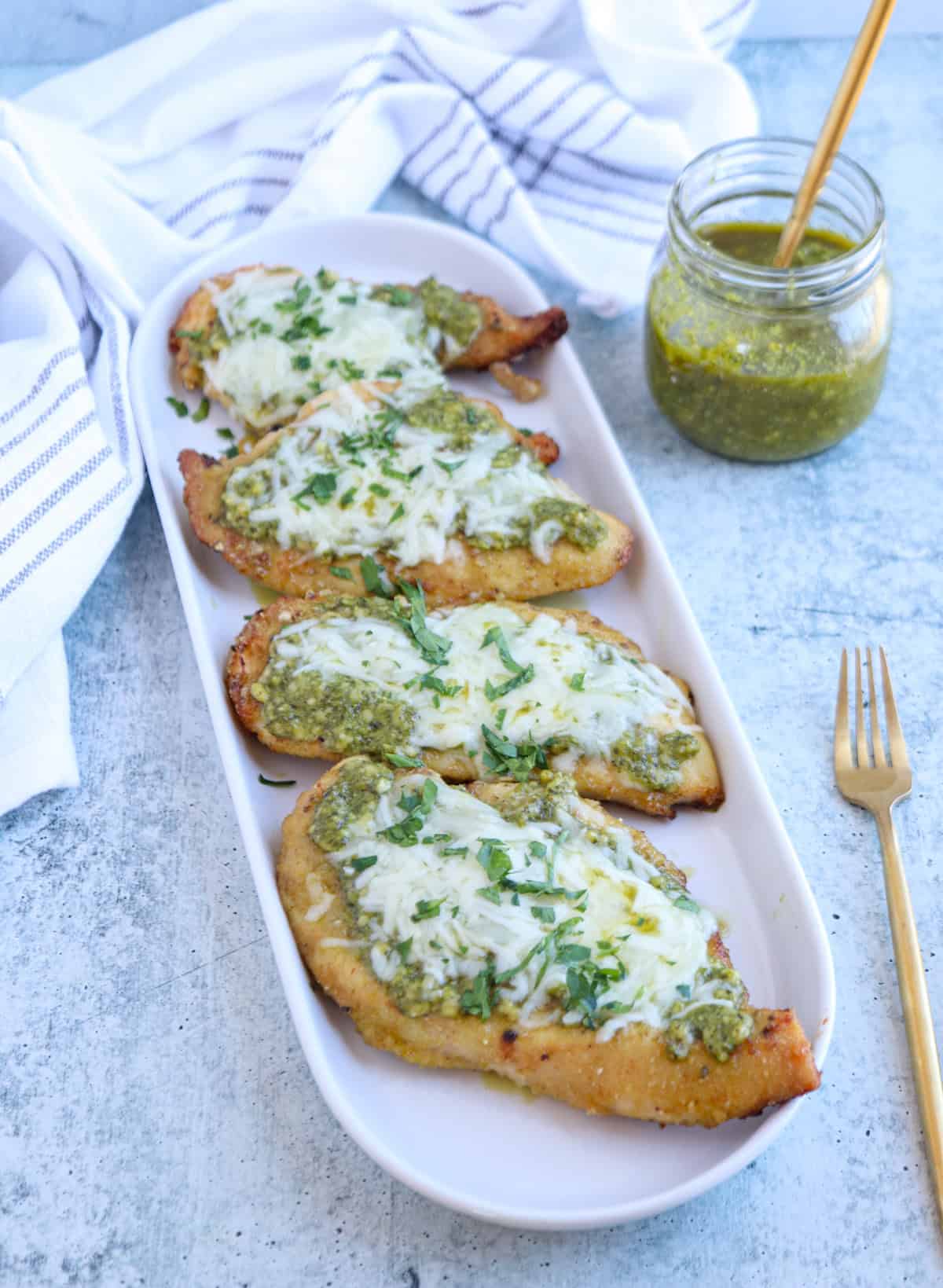 Four pieces of baked pesto chicken parmesan breast topped with melted cheese and green pesto, garnished with chopped herbs, served on a white oval plate. A jar of pesto and a gold fork are beside the plate.