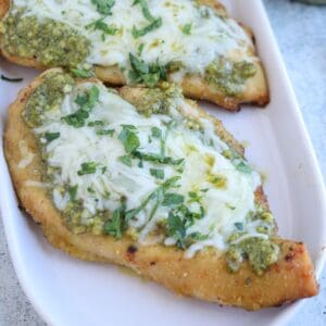Two pieces of baked pesto chicken parmesan, topped with melted cheese, green pesto sauce, and chopped fresh herbs, are served on a white rectangular dish.