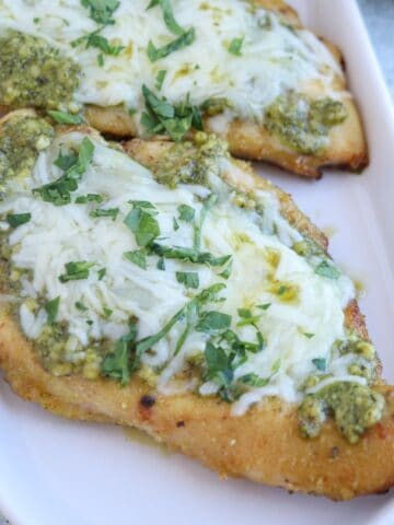 Two pieces of baked pesto chicken parmesan, topped with melted cheese, green pesto sauce, and chopped fresh herbs, are served on a white rectangular dish.