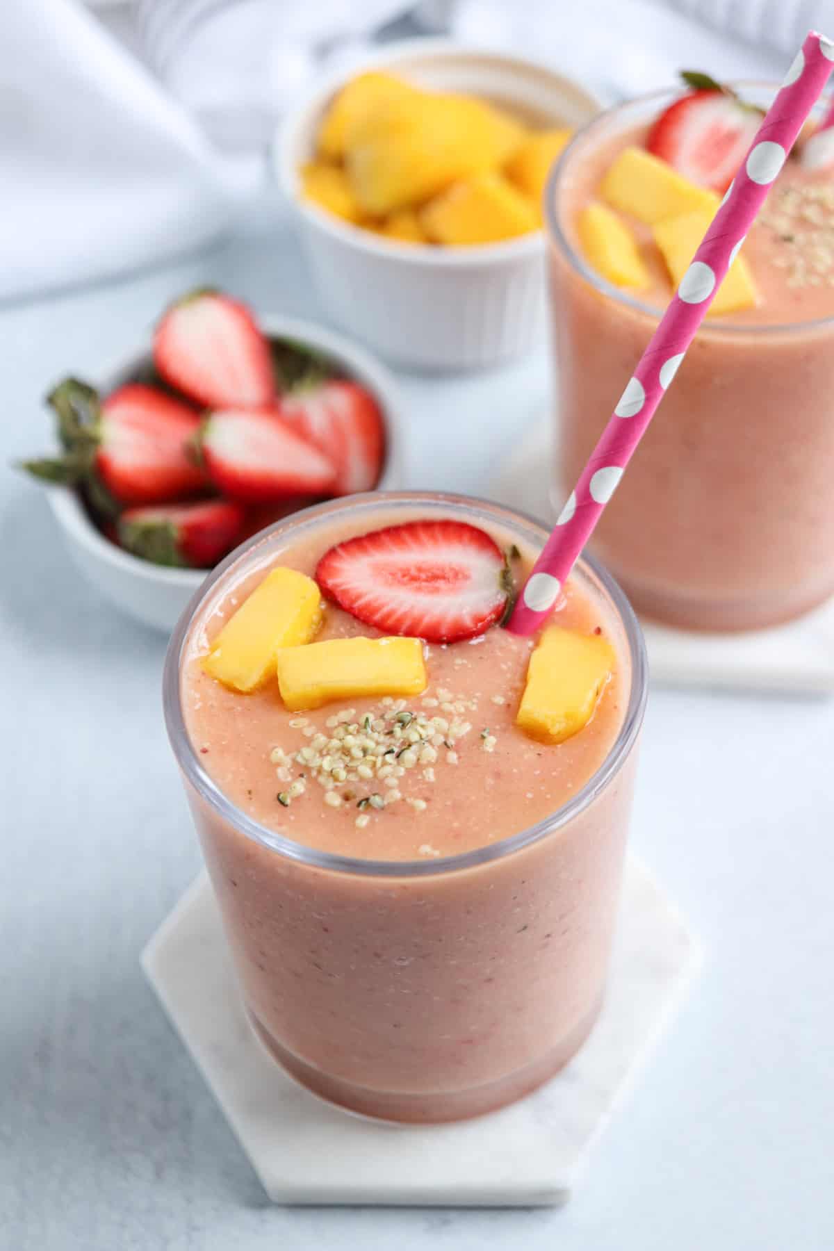 A glass of pink smoothie topped with mango chunks, a strawberry slice, and seeds, with a pink polka dot straw. In the background are bowls of cut strawberries, mango pieces, and a plate of pesto chicken parmesan.