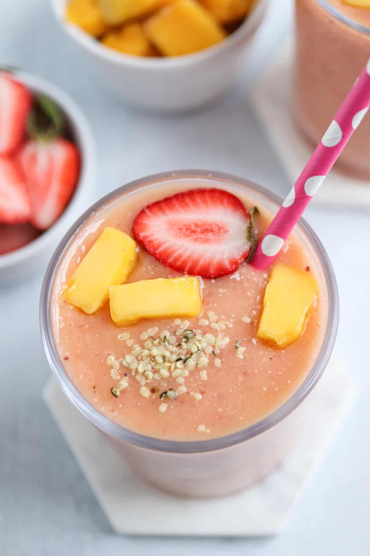 A glass of pink smoothie topped with a strawberry slice, mango pieces, and sprinkled seeds, with a pink polka-dot straw—perfect alongside a fresh pesto chicken parmesan. Bowls of sliced strawberries and mango are in the background.