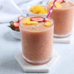 A glass of peach-colored smoothie topped with sliced strawberries, mango pieces, and seeds, with a pink polka-dotted straw, sits on a white coaster. In the background are another glass and a bowl of fruit—perfect after pesto chicken parmesan.