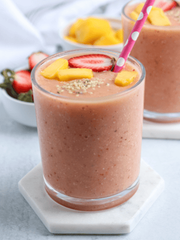 A glass of peach-colored smoothie topped with sliced strawberries, mango pieces, and seeds, with a pink polka-dotted straw, sits on a white coaster. In the background are another glass and a bowl of fruit—perfect after pesto chicken parmesan.