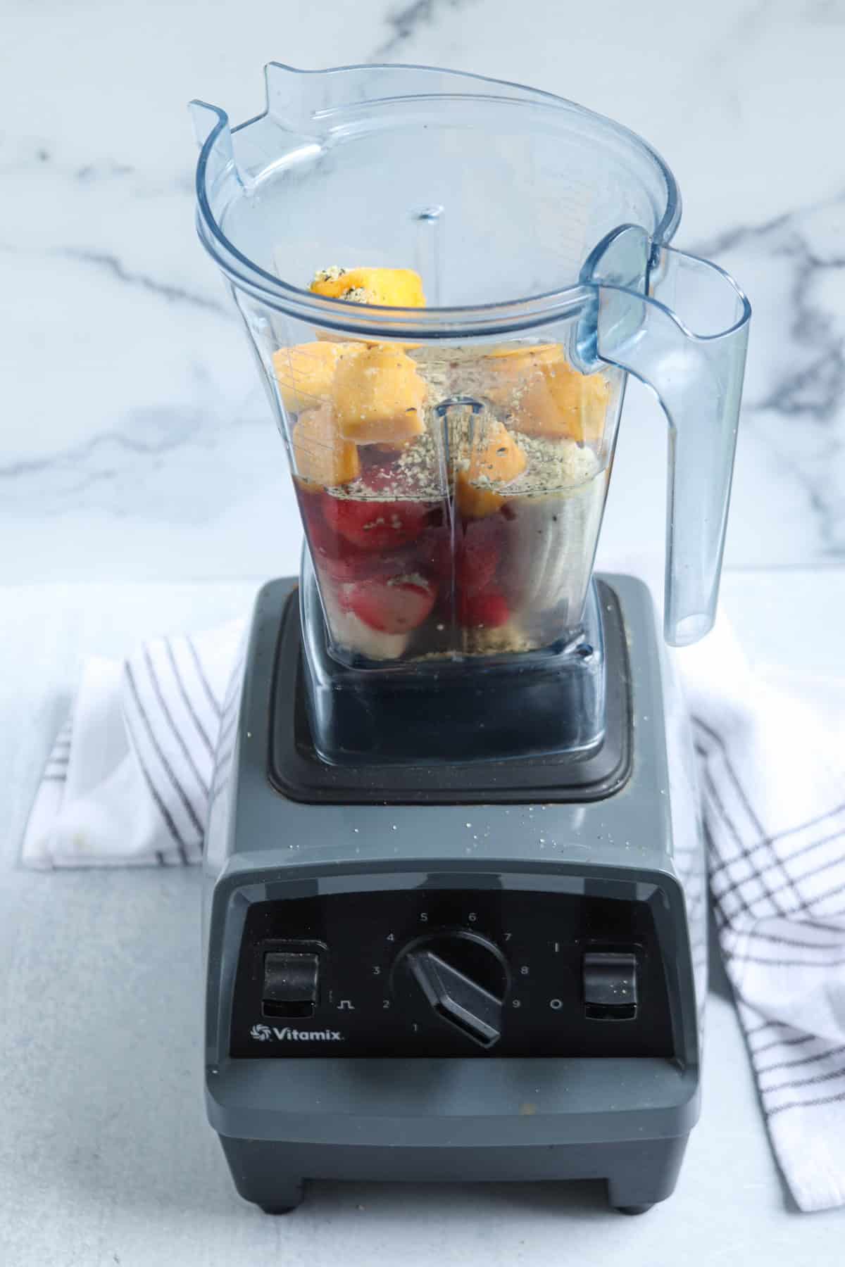 A blender filled with mango, strawberries, banana, and chia or hemp seeds sits on a white surface, ready to become a delicious strawberry mango banana smoothie. A white and gray striped towel is in the background.