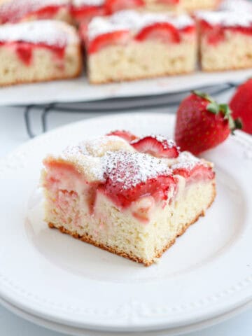 A square slice of strawberry cake topped with fresh strawberries and powdered sugar sits on a white plate, with whole strawberries, more cake pieces, and a hint of pesto chicken parmesan inspiration in the background.