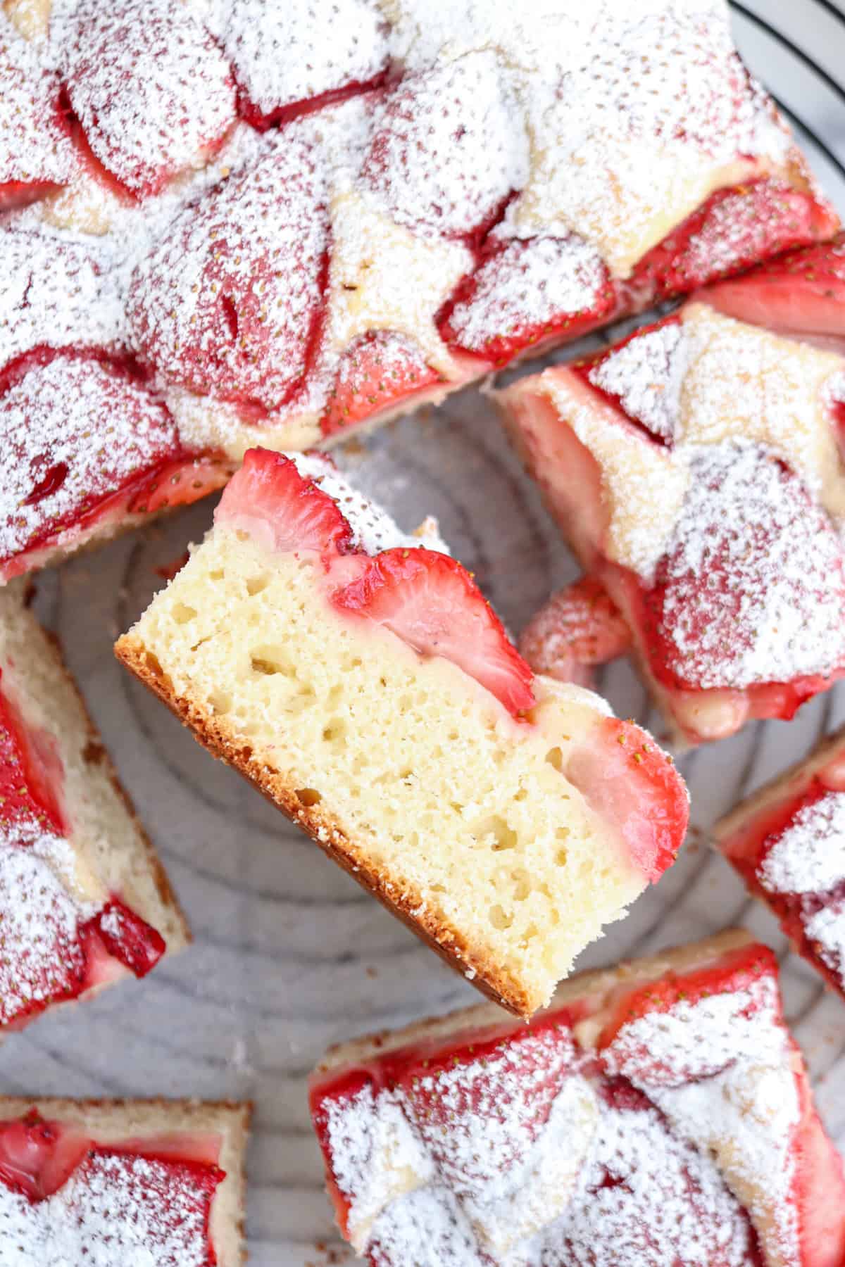 A slice of strawberry cake topped with powdered sugar is held up, showing its moist, fluffy texture—while more pieces are arranged on a plate in the background, perfect for enjoying after a savory pesto chicken parmesan meal.