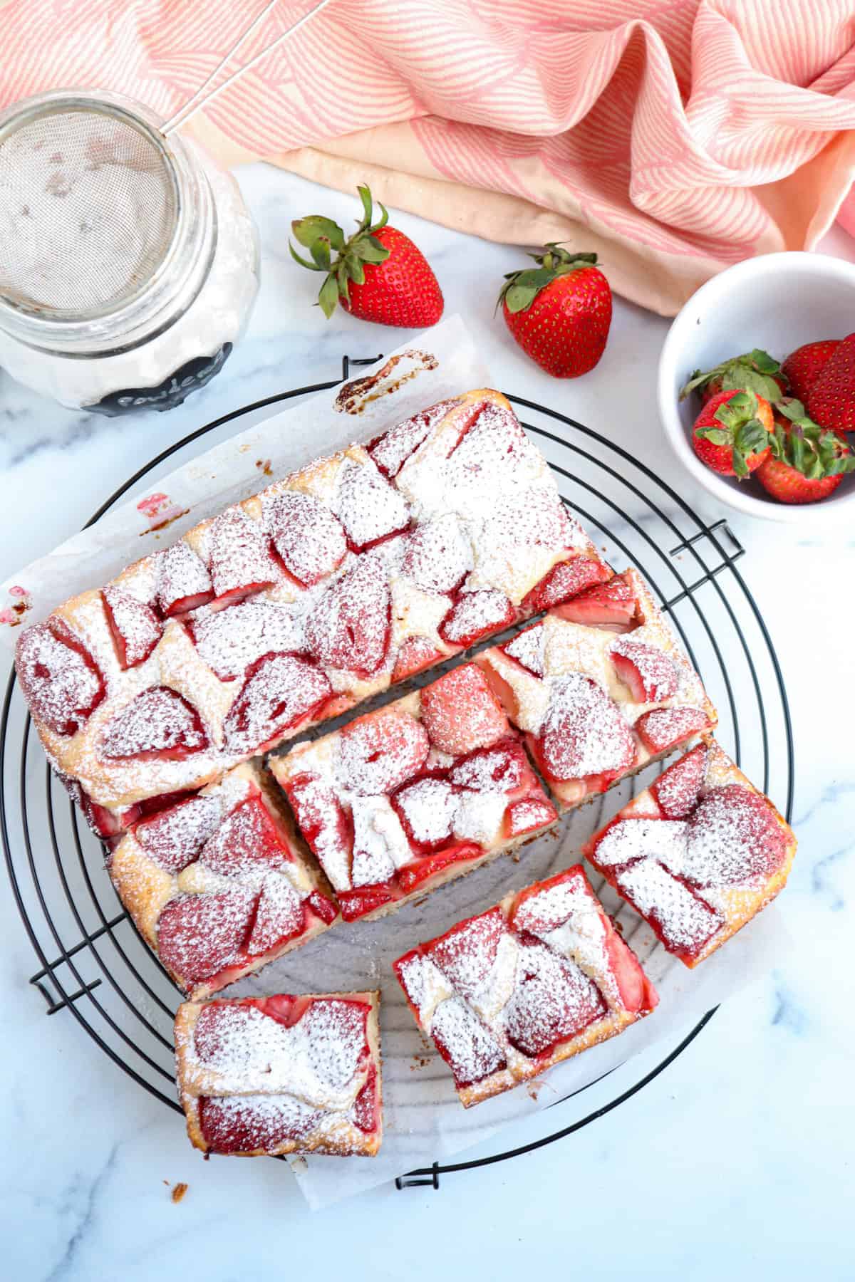 Rectangular strawberry cake dusted with powdered sugar, sliced into squares on a black wire rack. Nearby are fresh strawberries and a bowl with strawberry tops, creating a sweet scene—quite different from savory pesto chicken parmesan delights.