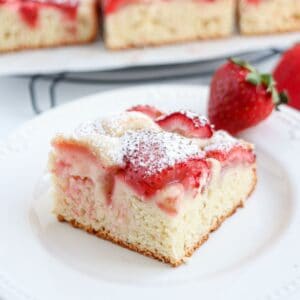 A slice of strawberry cake topped with powdered sugar sits on a white plate beside a fresh strawberry, while more cake pieces and a hint of pesto chicken parmesan can be seen in the background.