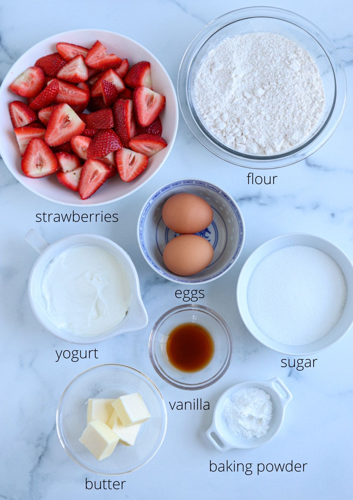 Bowls of sliced strawberries, flour, eggs, sugar, yogurt, butter, vanilla, and baking powder are arranged on a marble surface—much like prepping for a pesto chicken parmesan recipe—each labeled with its ingredient name.