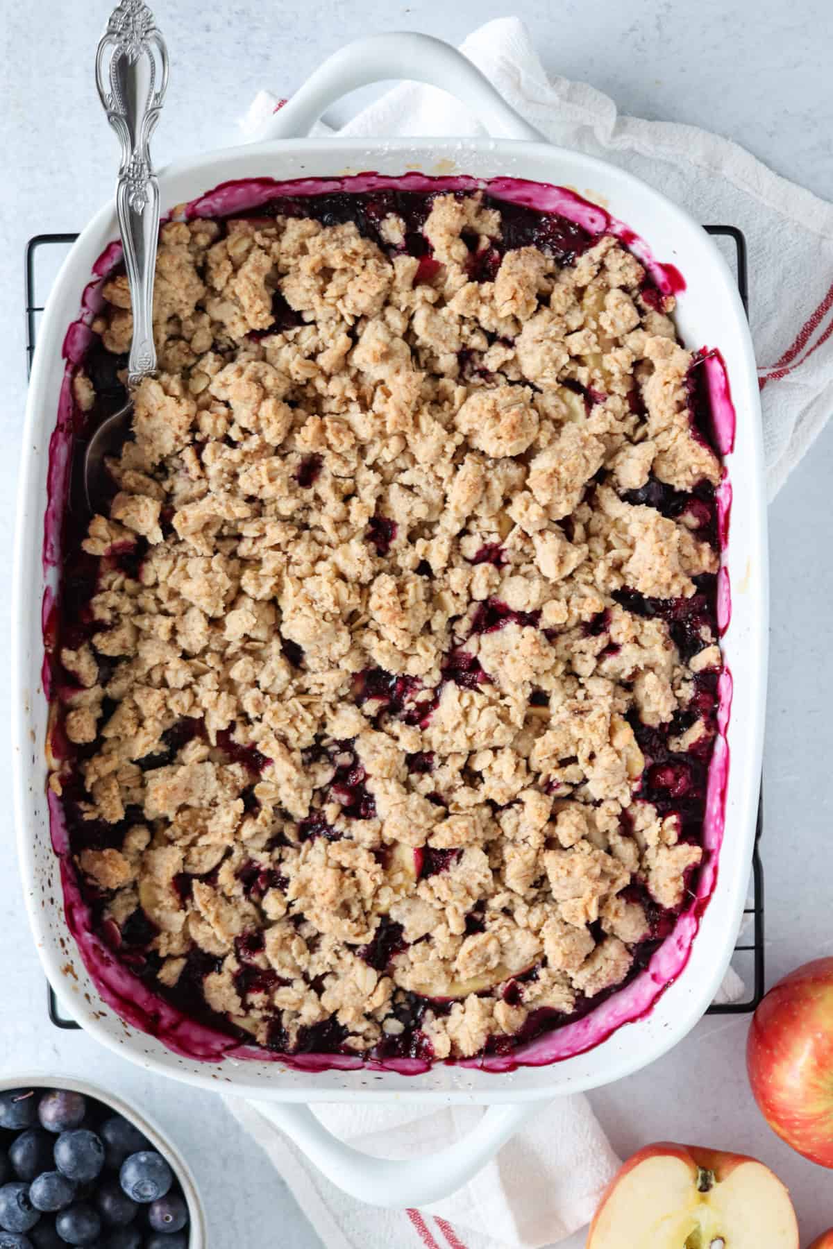 A baked fruit crumble dessert in a white rectangular dish with a serving spoon, showing a golden crumb topping and berry filling. The dish rests on a white towel, with blueberries, an apple, and hints of pesto chicken parmesan nearby.