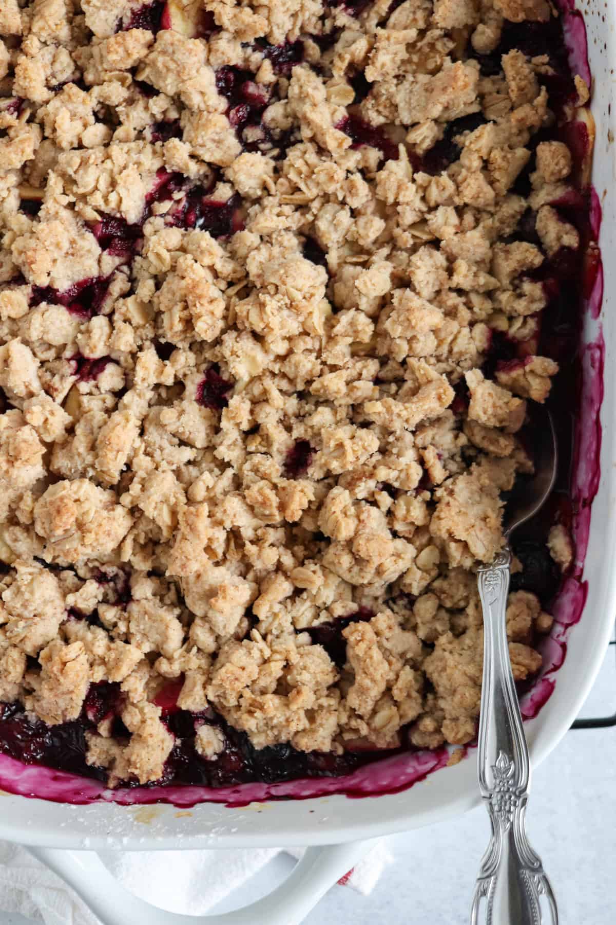 A close-up of a baked fruit crumble in a white dish, topped with a golden-brown oat and crumb topping. A silver spoon rests in the dish, similar to how you might serve pesto chicken parmesan, revealing dark berry filling underneath.