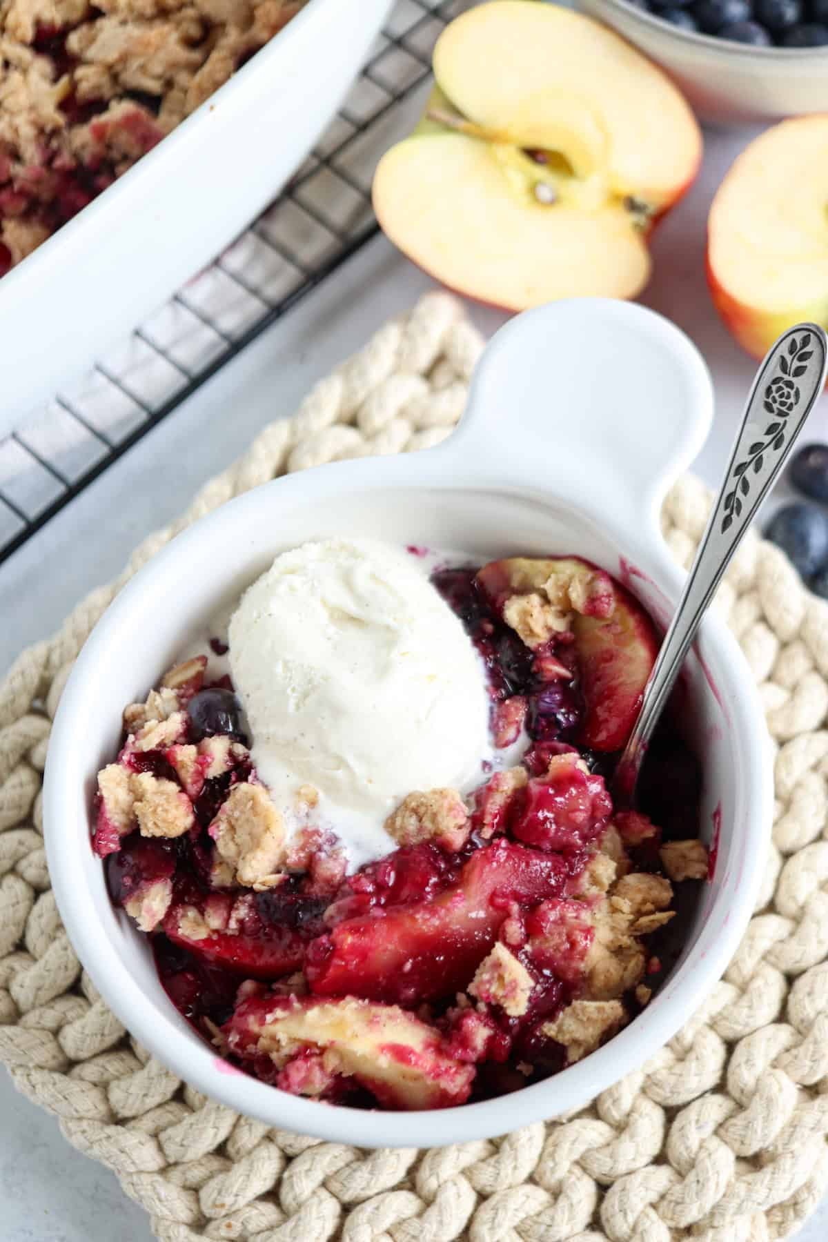 A bowl of fruit crumble with peaches and berries, topped with a scoop of vanilla ice cream. A spoon rests in the bowl, and sliced peaches and blueberries are visible in the background—a sweet treat after pesto chicken parmesan.