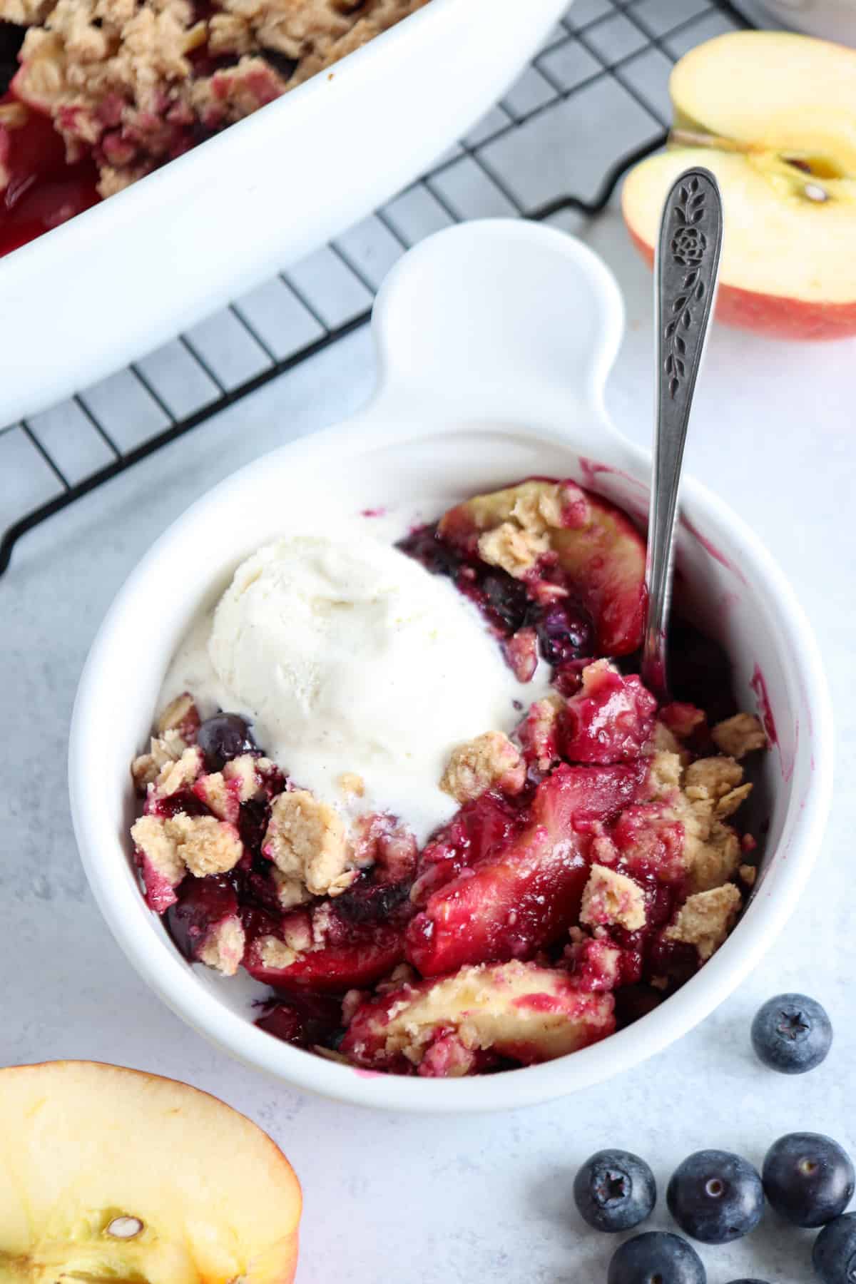 A white bowl filled with fruit crumble made of peaches and blueberries, topped with a scoop of vanilla ice cream. A spoon is in the bowl, and fresh peach slices and blueberries are nearby—perfect after savoring pesto chicken parmesan.