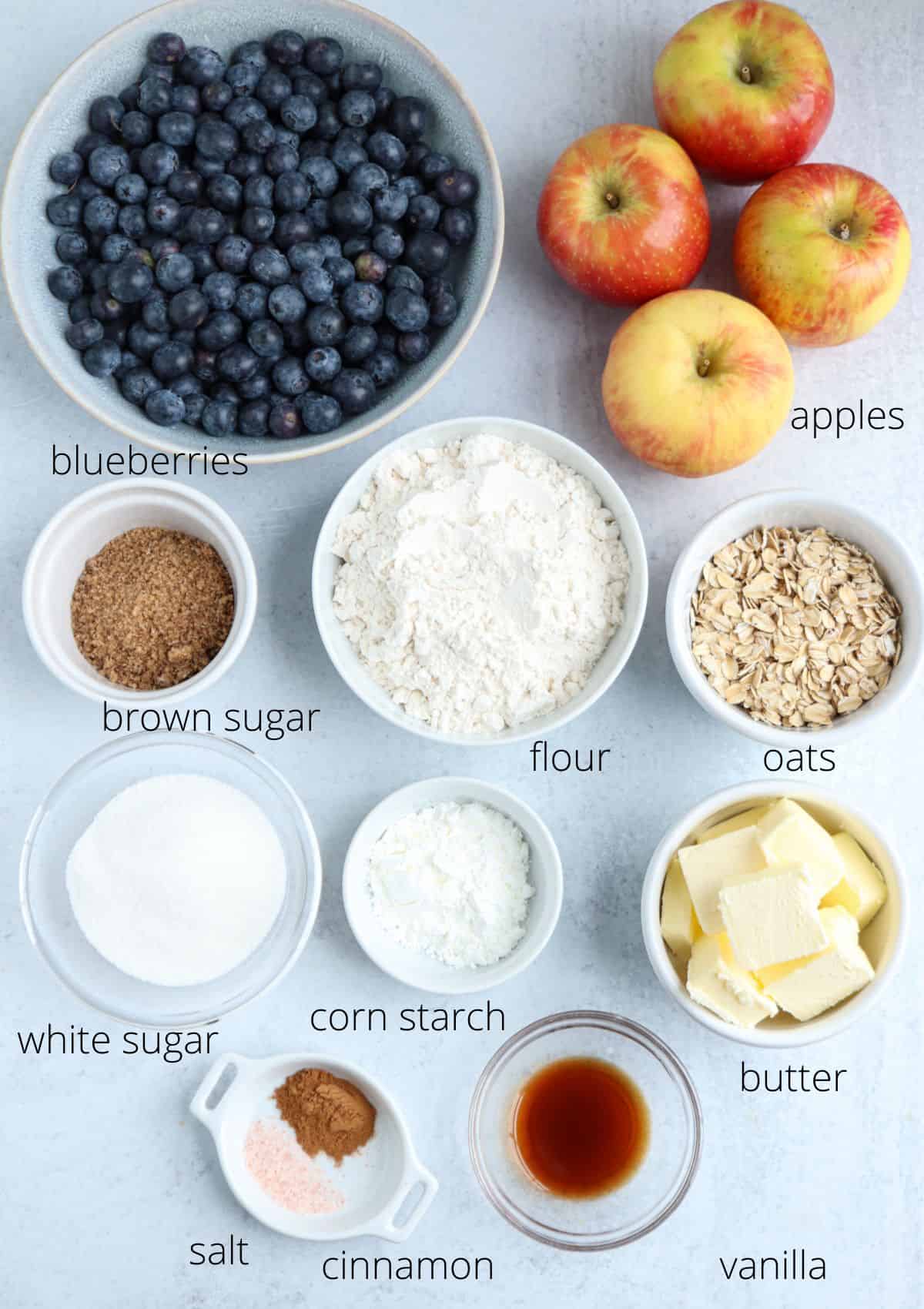 Top-down view of labeled baking ingredients for a sweet treat—blueberries, apples, brown sugar, flour, oats, and more—in separate bowls on a light surface. Not intended for pesto chicken parmesan recipes.