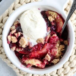 A bowl of fruit crumble with slices of baked fruit and berries, topped with a scoop of vanilla ice cream, served in a white dish beside pesto chicken parmesan on a textured, beige mat.