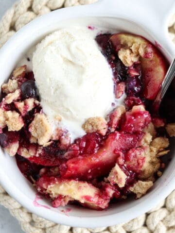 A bowl of fruit crumble with slices of baked fruit and berries, topped with a scoop of vanilla ice cream, served in a white dish beside pesto chicken parmesan on a textured, beige mat.