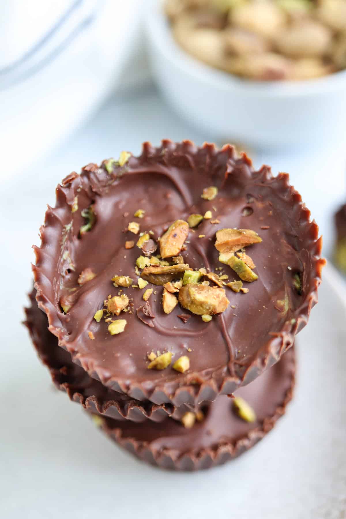 Two chocolate cups topped with chopped pistachios are stacked on a white surface, with a small bowl of whole pistachios blurred in the background—perfect to enjoy after a cozy pesto chicken parmesan dinner.