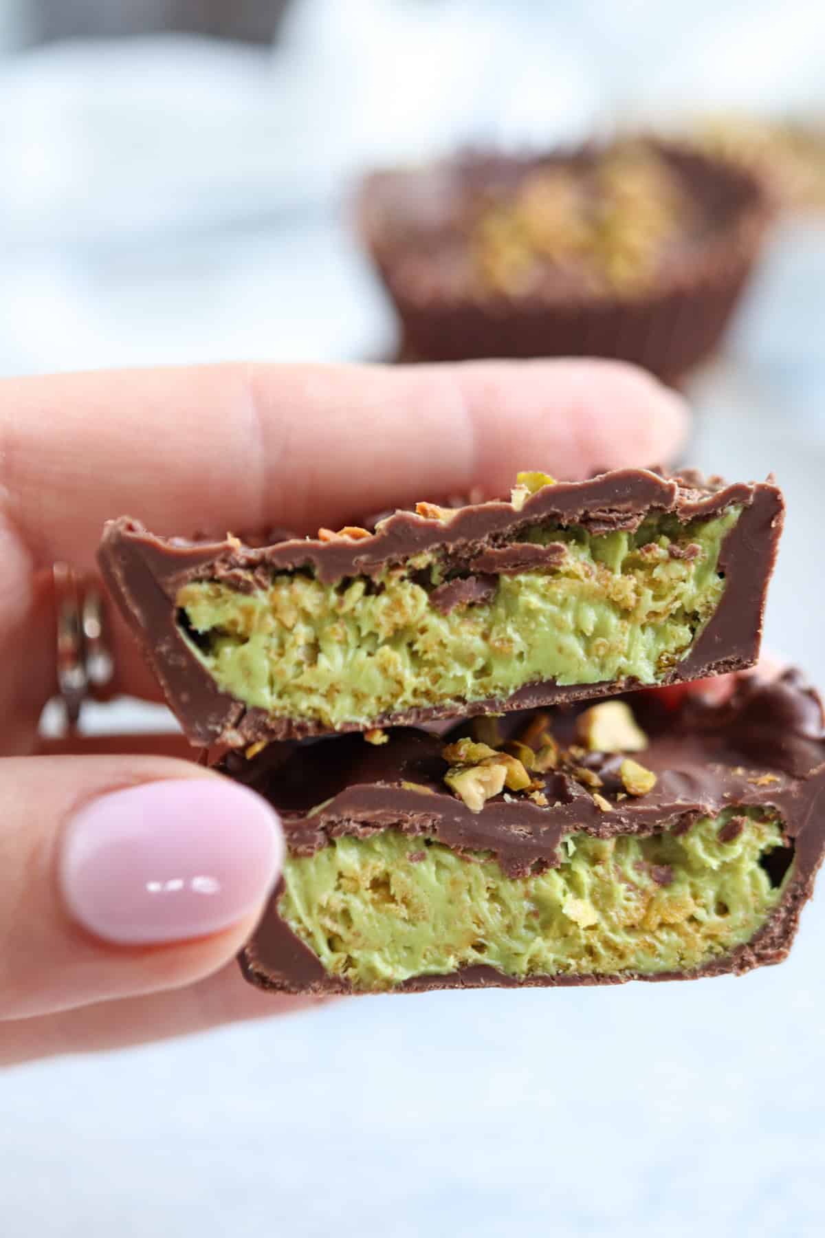 A hand with light pink nail polish holds two halves of a chocolate cup filled with a green pistachio mixture, reminiscent of the vibrant color in pesto chicken parmesan, and topped with crushed pistachios.
