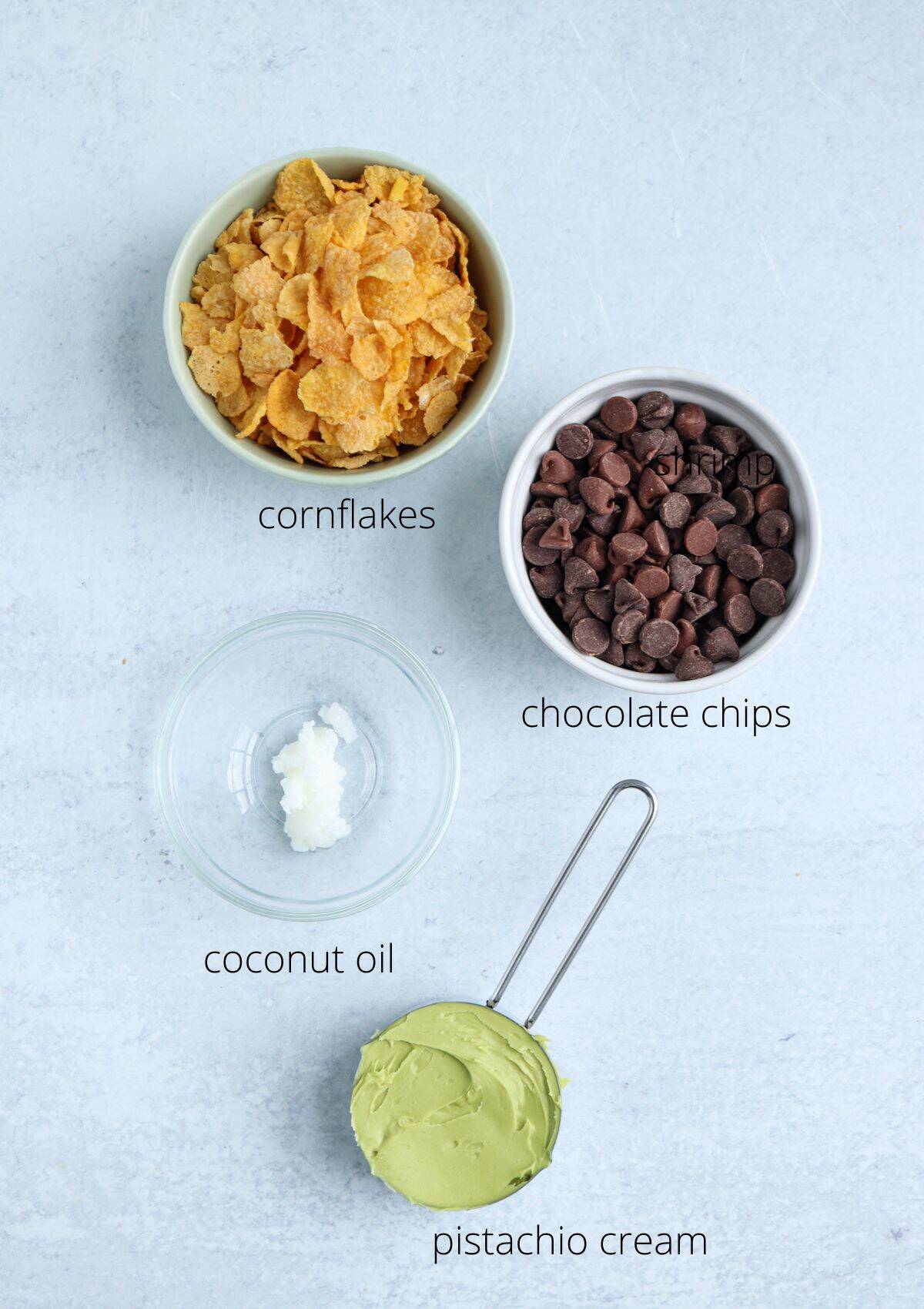 Four ingredients on a light surface: a bowl of cornflakes, a bowl of chocolate chips, coconut oil in a small glass dish, and pistachio cream in a metal measuring cup—perfect for adding to unique treats like pesto chicken parmesan.