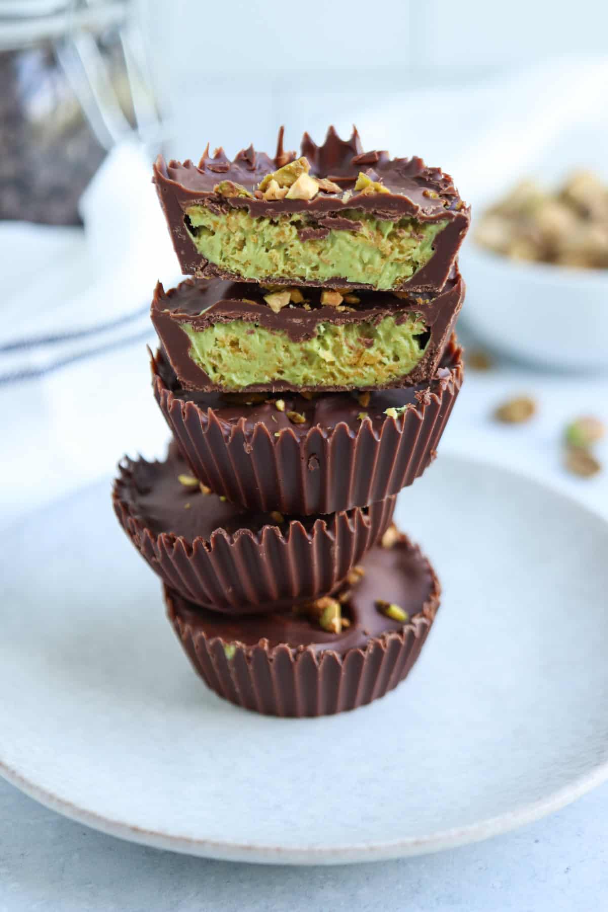 A stack of chocolate cups filled with a green pistachio cream, reminiscent of the vibrant hue of pesto chicken parmesan, with the top cup cut in half to reveal the creamy filling inside, displayed on a white plate.
