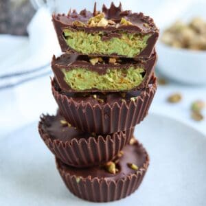 A stack of chocolate cups with a creamy green pistachio filling, reminiscent of pesto chicken parmesan hues, topped and sprinkled with chopped pistachios, on a light plate with a blurred background.