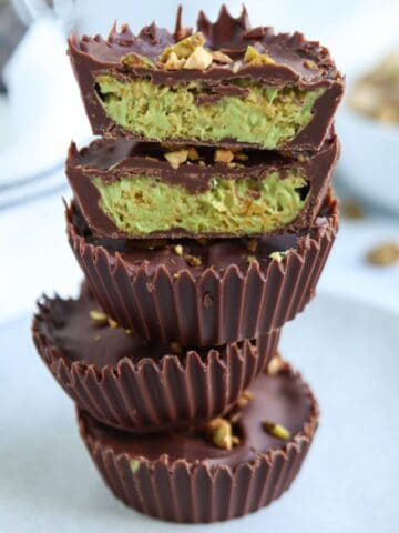 A stack of chocolate cups with a creamy green pistachio filling, reminiscent of pesto chicken parmesan hues, topped and sprinkled with chopped pistachios, on a light plate with a blurred background.