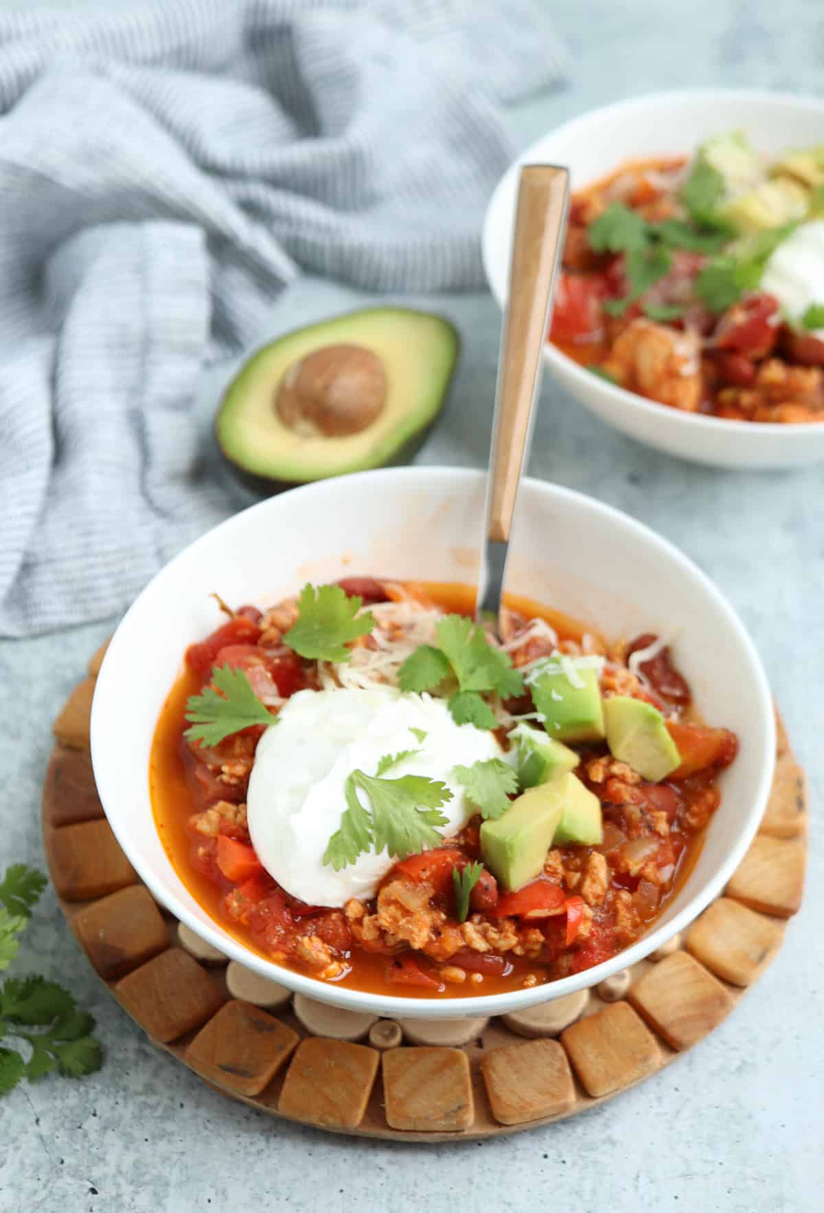 A bowl of chili topped with sour cream, shredded cheese, diced avocado, and cilantro. A spoon rests in the bowl, while a halved avocado, napkin, and a second chili or pesto chicken parmesan bowl sit nearby on a light surface.