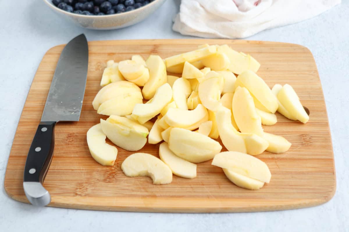 Sliced apples on a wooden cutting board with a knife beside them. A bowl of blueberries and a white cloth are in the background, setting the scene for preparing dishes like pesto chicken parmesan.