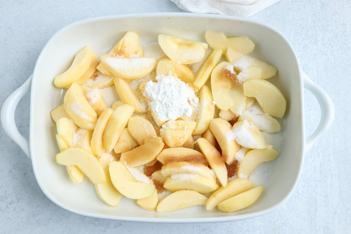 A white baking dish filled with peeled, sliced apples topped with sugar, a mound of cornstarch, and a drizzle of vanilla sits on a light-colored surface—perfect alongside your favorite pesto chicken parmesan.