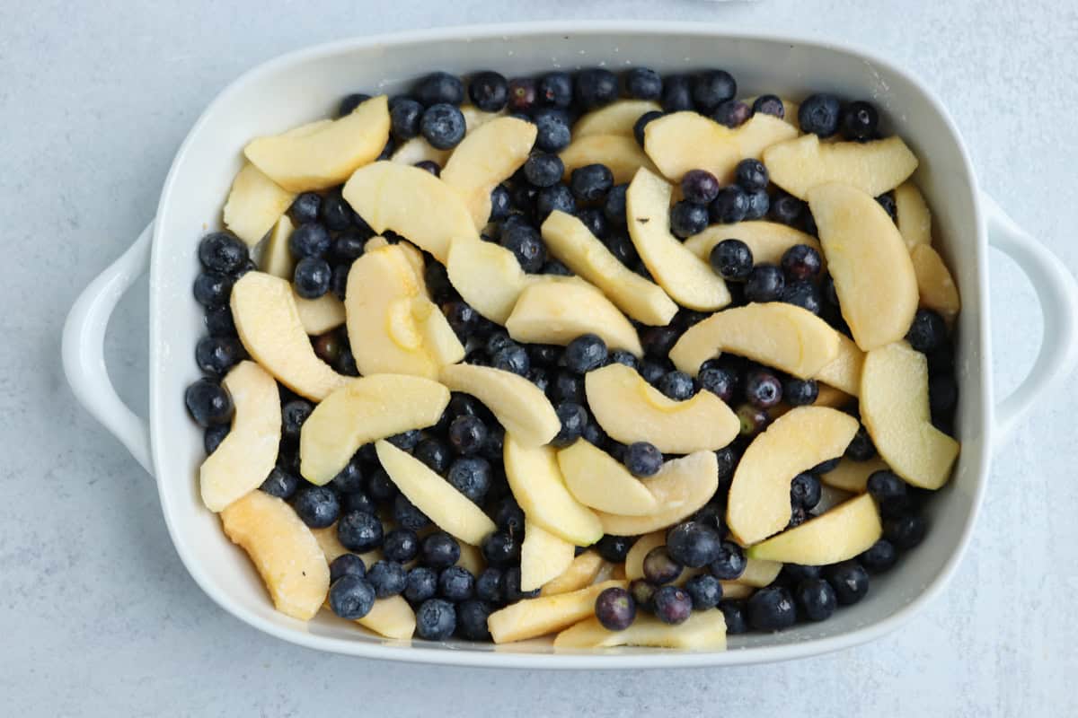 A white baking dish filled with sliced apples and whole blueberries, uncooked, sits on a light gray surface—perfect for dessert or to serve after enjoying pesto chicken parmesan.