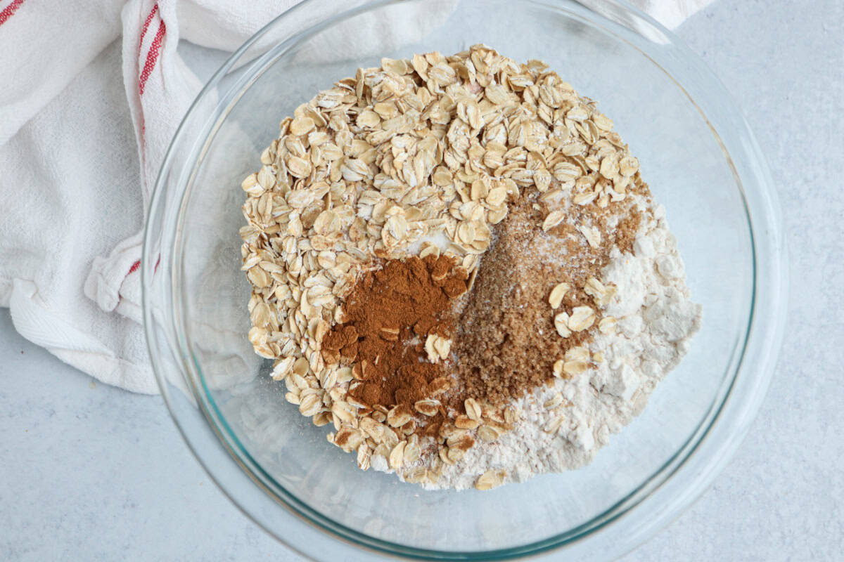 A glass bowl filled with dry baking ingredients including rolled oats, brown sugar, flour, and ground cinnamon sits on a light surface next to a white kitchen towel with red stripes—perfect prep for your next pesto chicken parmesan bake.
