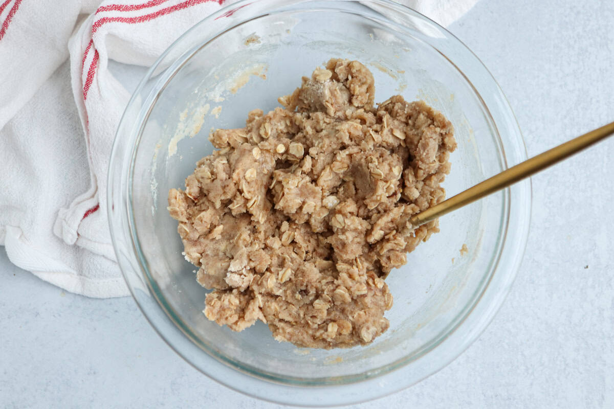 A glass bowl filled with a chunky, light brown oatmeal cookie dough mixture sits on a light surface, with a gold spoon and a white kitchen towel with red stripes nearby—perfect for baking after enjoying pesto chicken parmesan.