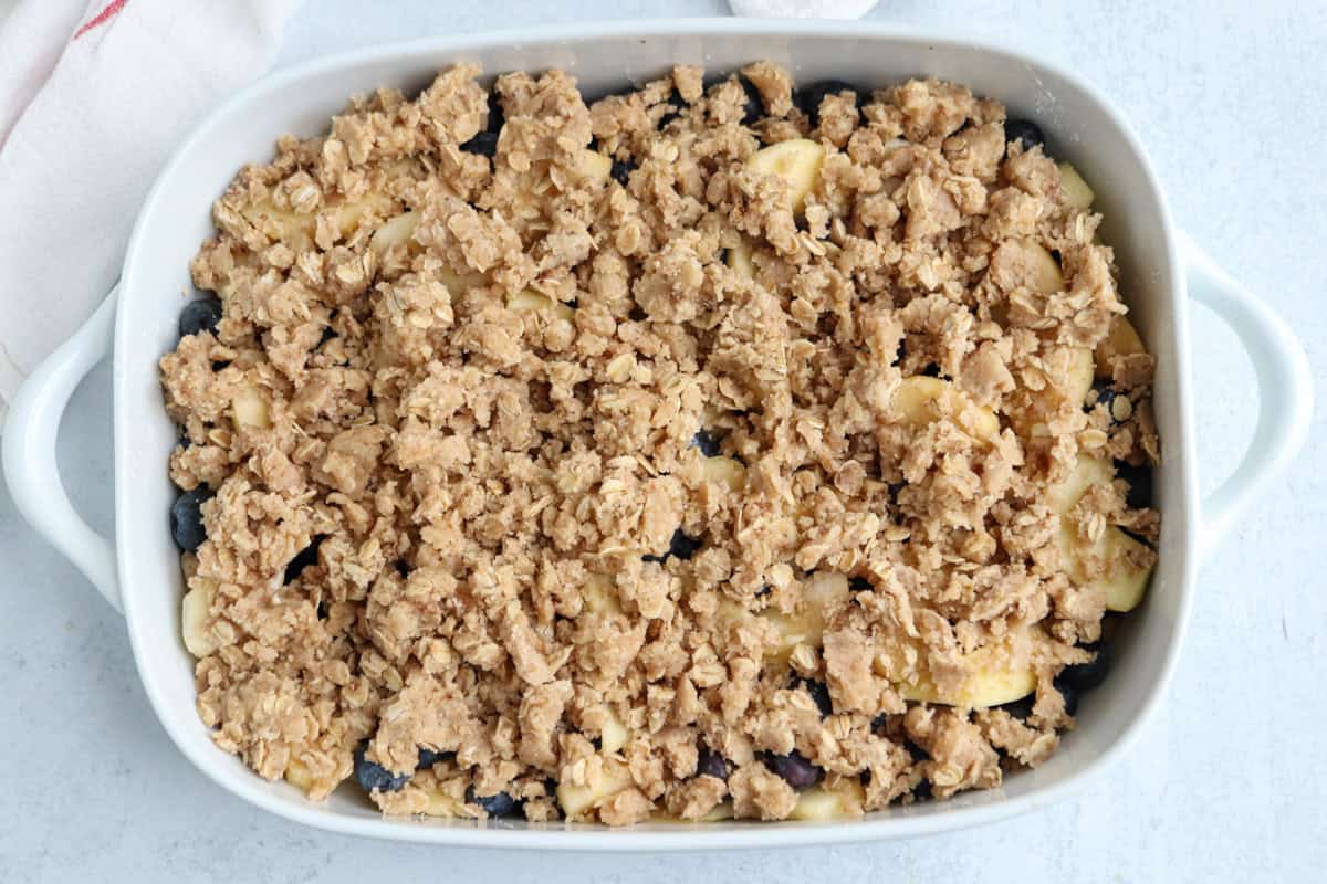 A white baking dish filled with an unbaked fruit crisp, showing a layer of sliced fruit and berries topped with a crumbly oat and brown sugar topping, sits beside a bubbling pesto chicken parmesan, both ready for the oven.