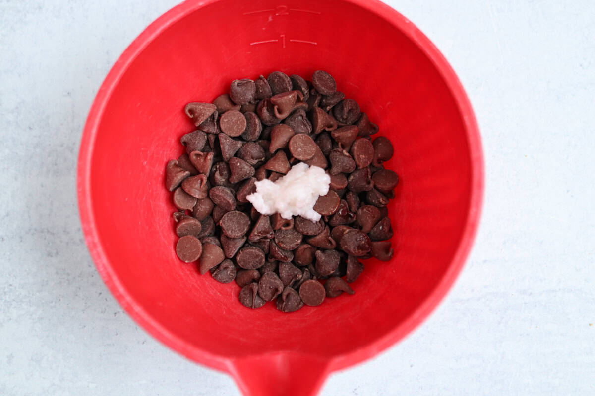 A red mixing bowl filled with chocolate chips and a dollop of white coconut oil in the center, placed on a light gray surface—ready for baking or pairing sweetly after a savory pesto chicken parmesan dinner.