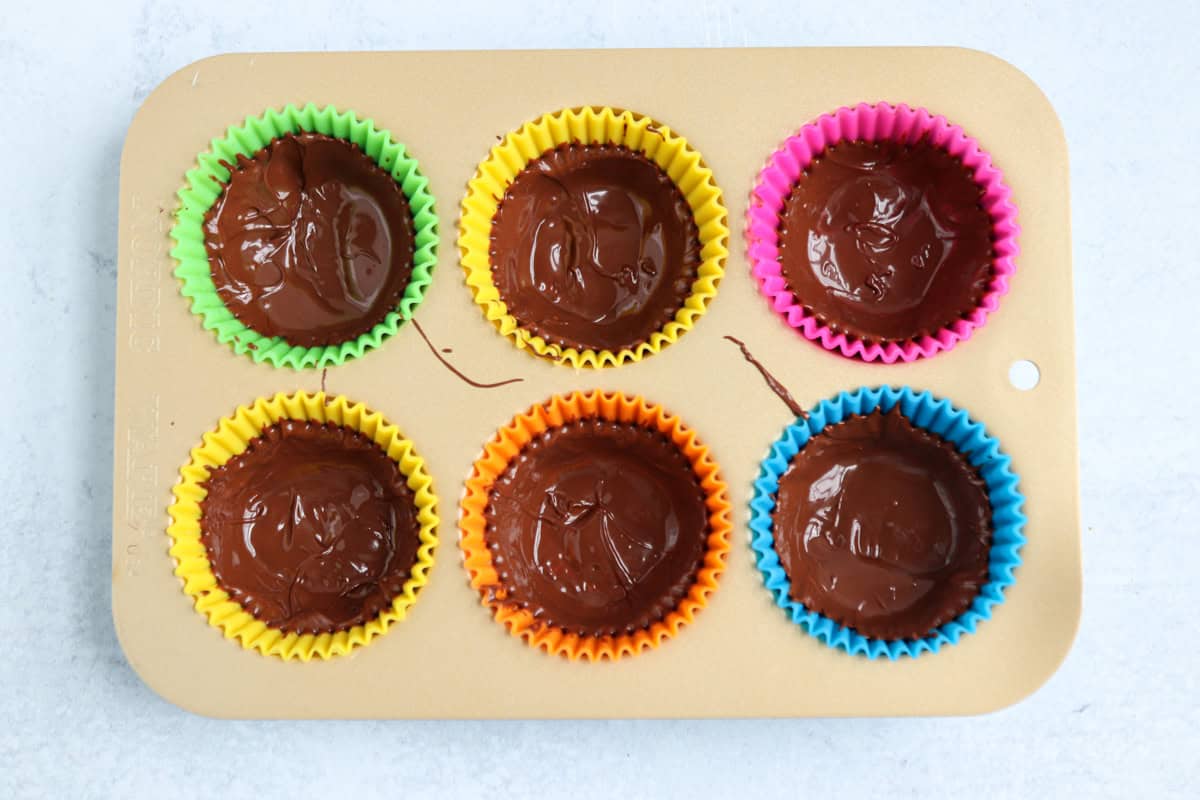 A muffin tin lined with colorful cupcake wrappers, each filled with melted chocolate, sits on a light surface—perfect for a sweet treat or serving alongside savory favorites like pesto chicken parmesan.