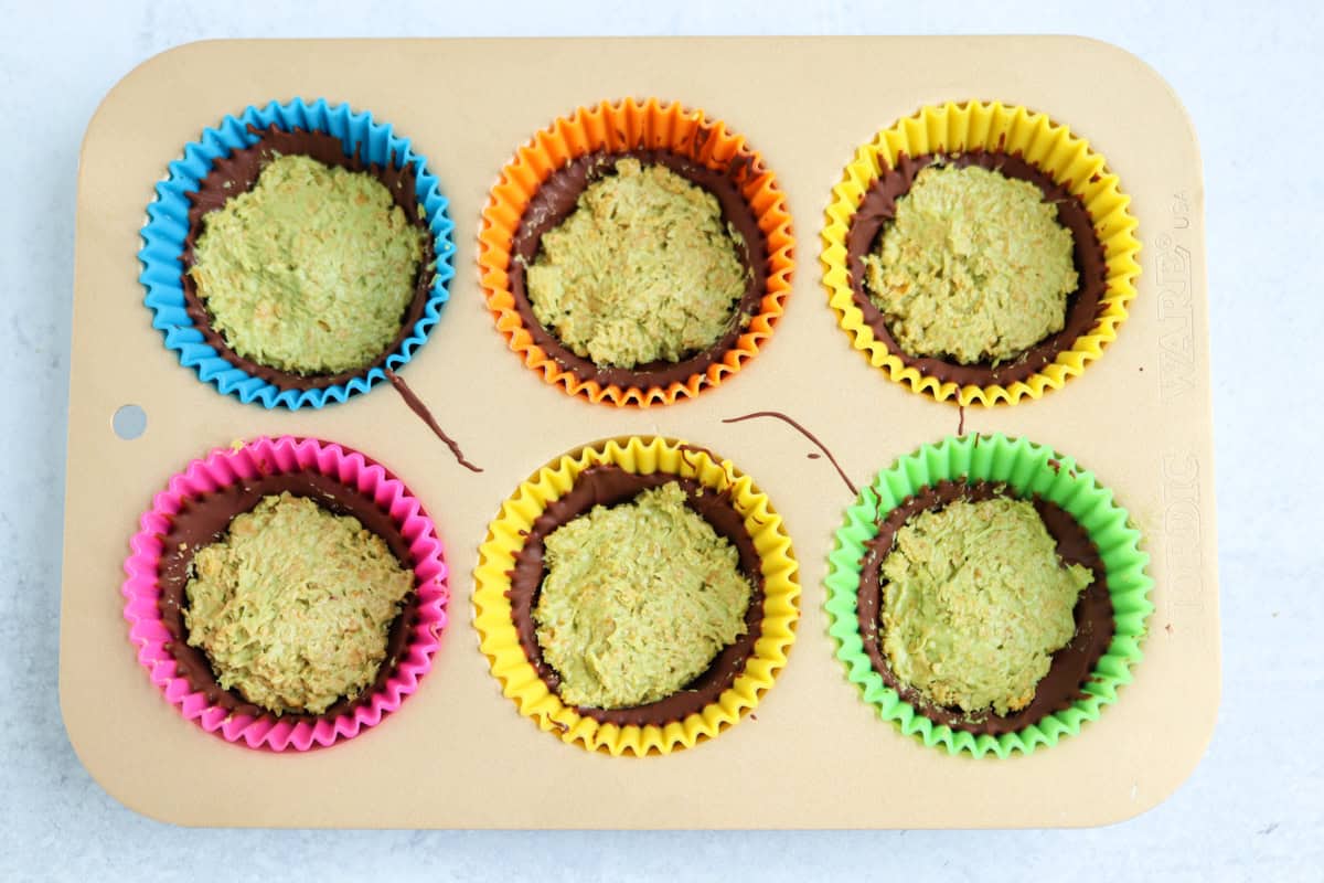 A tan muffin tray holds six colorful silicone cupcake liners, each filled with a scoop of unbaked, pesto chicken parmesan-inspired greenish dough atop a chocolate layer. The liners are pink, blue, orange, green, and two yellow.