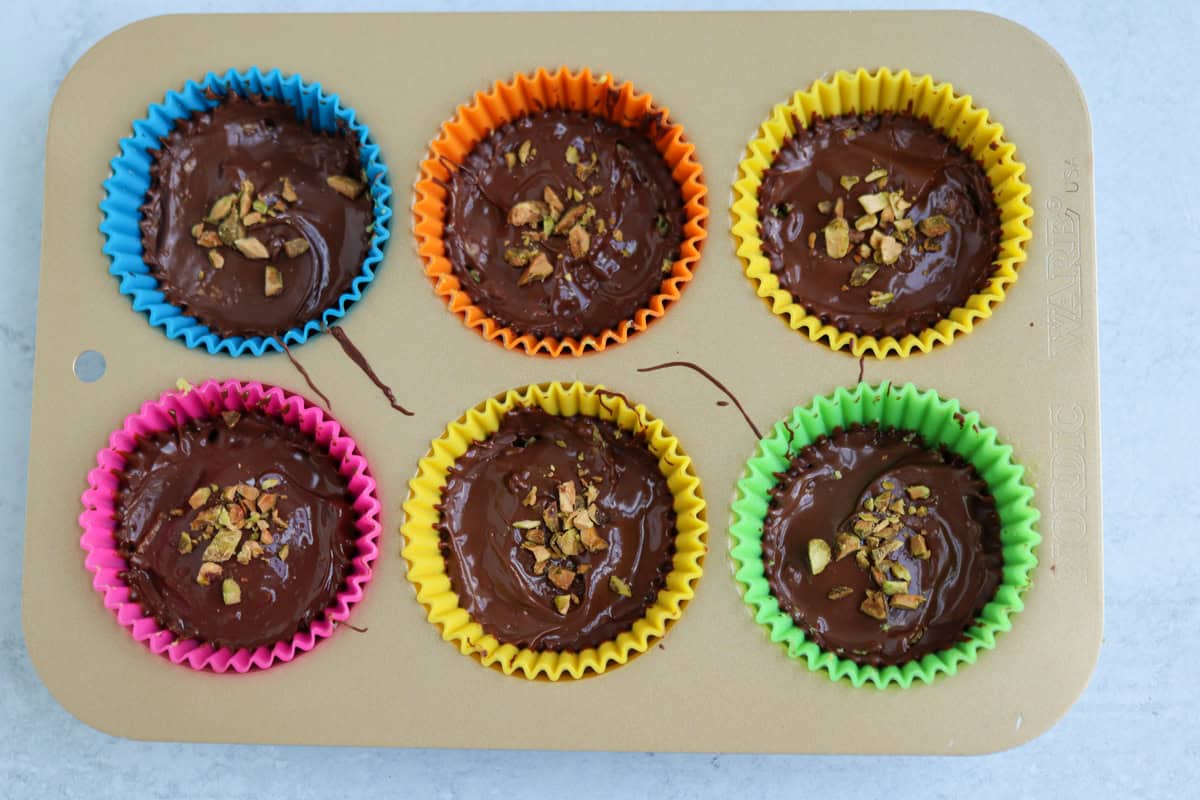 A muffin tray with six colorful silicone liners, each filled with melted chocolate and topped with chopped nuts—perfect to enjoy after a delicious pesto chicken parmesan meal, ready to be baked or set.
