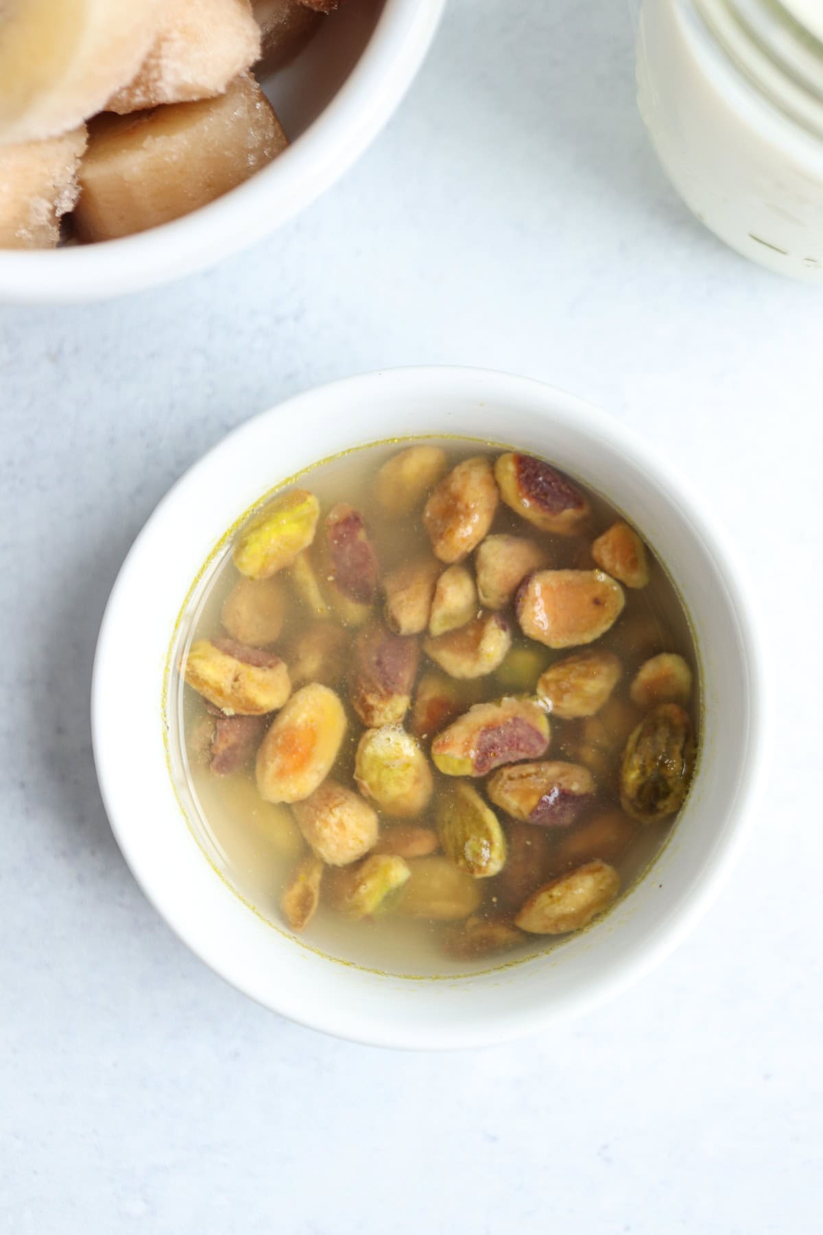 A white bowl filled with pistachios soaking in water sits on a light surface, ready for a creamy pistachio smoothie. Nearby, part of a bowl with frozen cubes and the edge of a jar are partially visible.