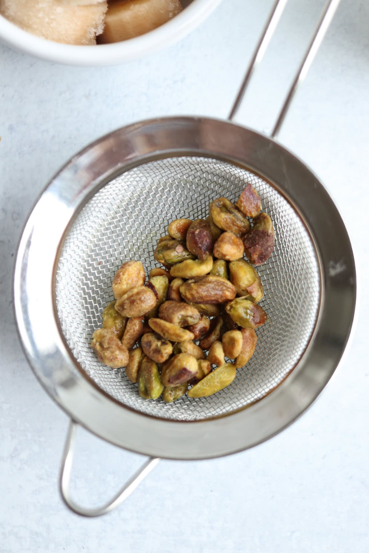 A metal strainer with shelled pistachios inside sits on a light surface. In the corner, a partial white bowl with brown contents hints at ingredients for a creamy pistachio smoothie.