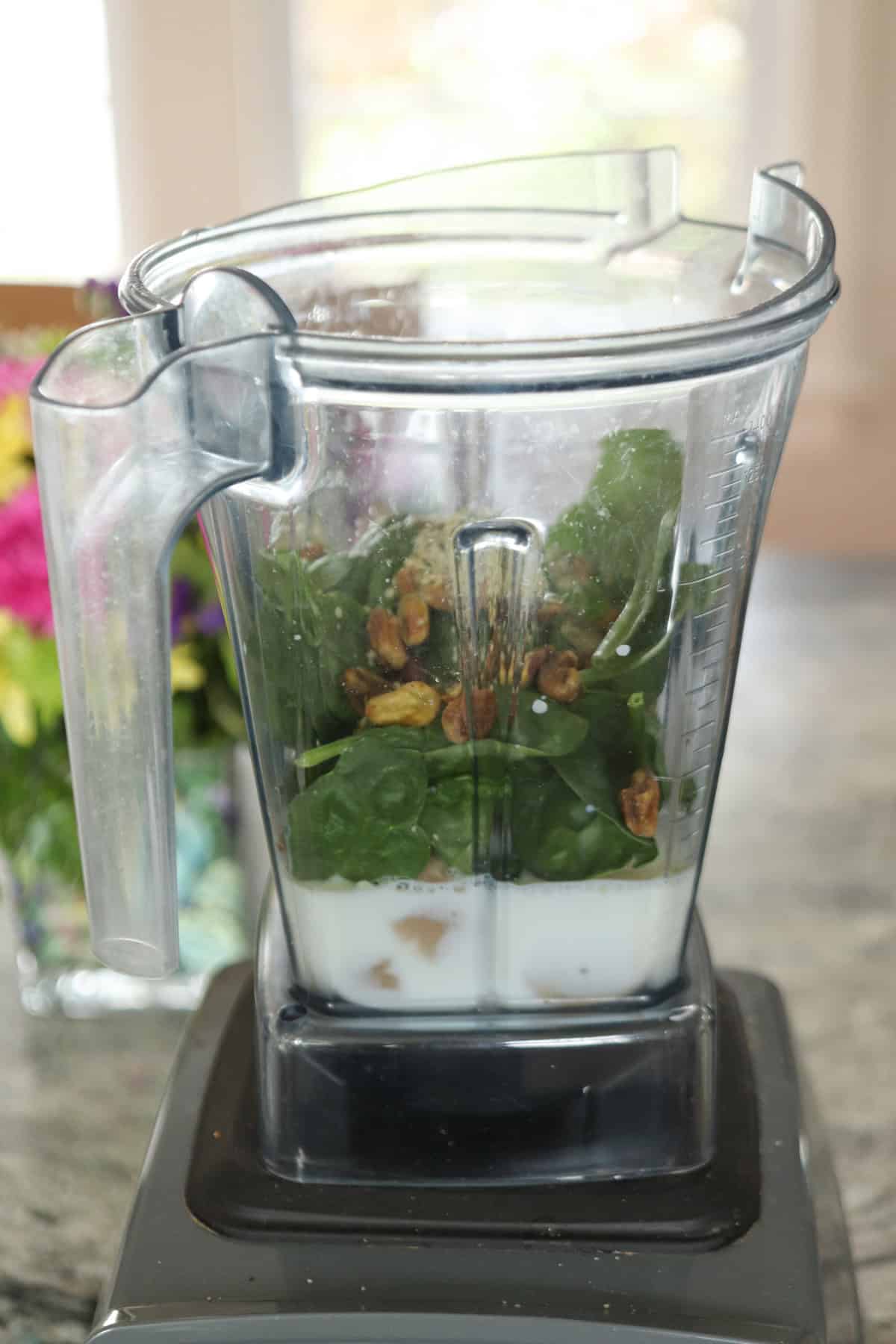 A blender jar filled with spinach leaves, nuts, and a white liquid, likely milk or yogurt, sits on a kitchen countertop—perfect ingredients for a refreshing pistachio smoothie.