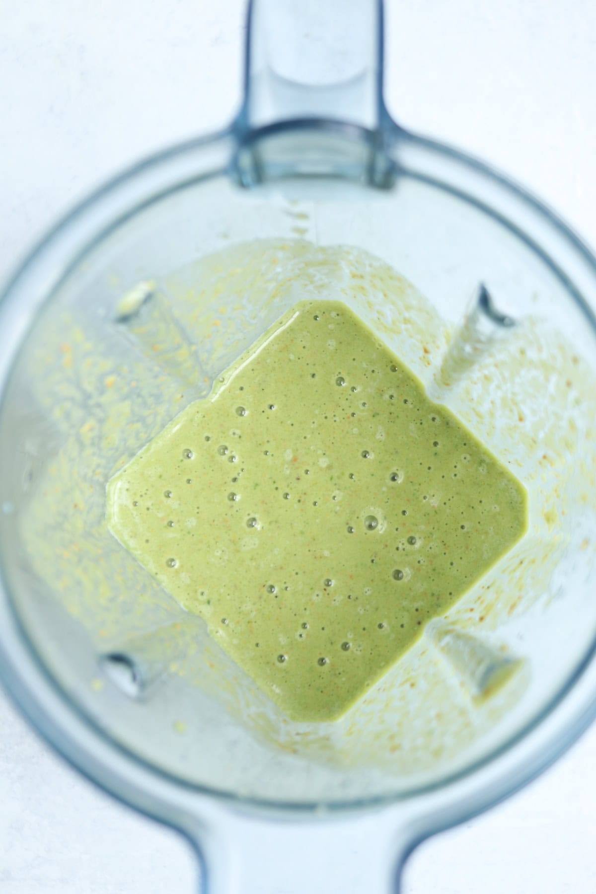 A top-down view of a blender containing a thick, light green pistachio smoothie with small bubbles on the surface. The blender walls are coated with some of the mixture.