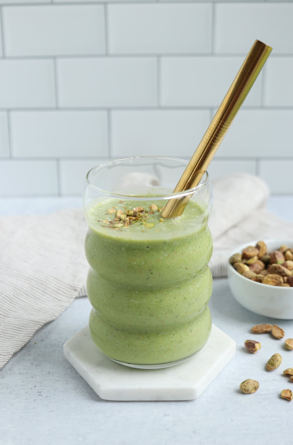 A pistachio smoothie in a wavy glass is garnished with chopped pistachios and a gold straw, set on a white coaster with a bowl of pistachios and a beige cloth in the background.
