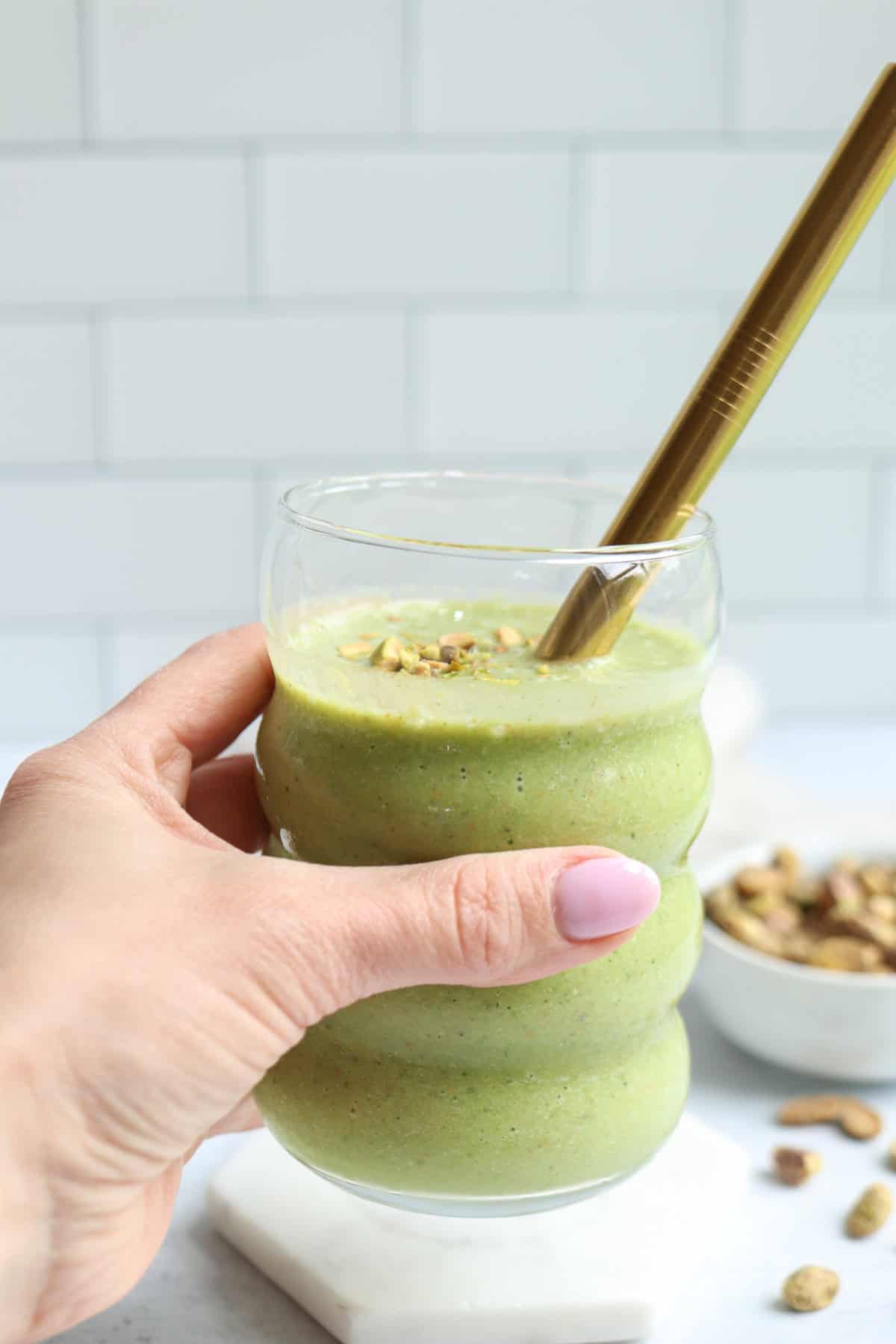 A hand with light pink nail polish holds a curvy glass filled with a green pistachio smoothie, topped with chopped nuts. A gold metal straw rests in the glass. In the blurred background, a bowl of nuts is visible.