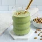 A glass of pistachio smoothie topped with chopped pistachios and a gold straw sits on a white hexagonal coaster, with a small bowl of pistachios and a beige cloth in the background.
