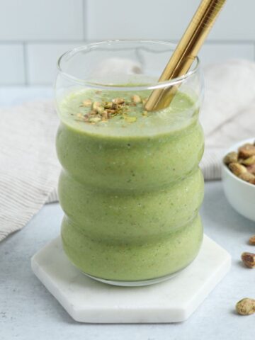 A glass of pistachio smoothie topped with chopped pistachios and a gold straw sits on a white hexagonal coaster, with a small bowl of pistachios and a beige cloth in the background.