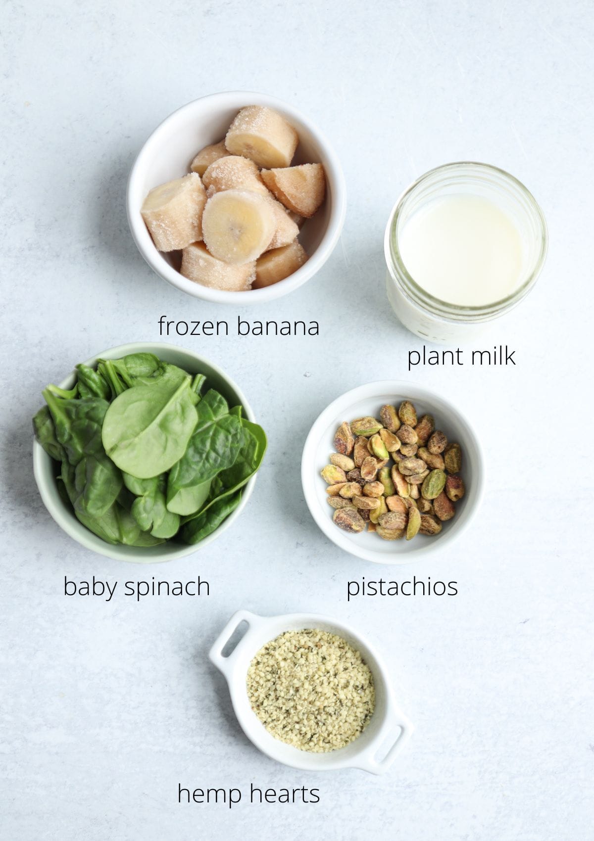 Five bowls on a white surface contain labeled ingredients for a pistachio smoothie: frozen banana slices, plant milk in a jar, baby spinach leaves, shelled pistachios, and hemp hearts.