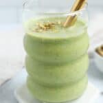 A clear, curvy glass filled with a thick pistachio smoothie, topped with chopped pistachios and a gold metal straw, rests on a white hexagonal coaster. Text above reads PISTACHIO SMOOTHIE.
