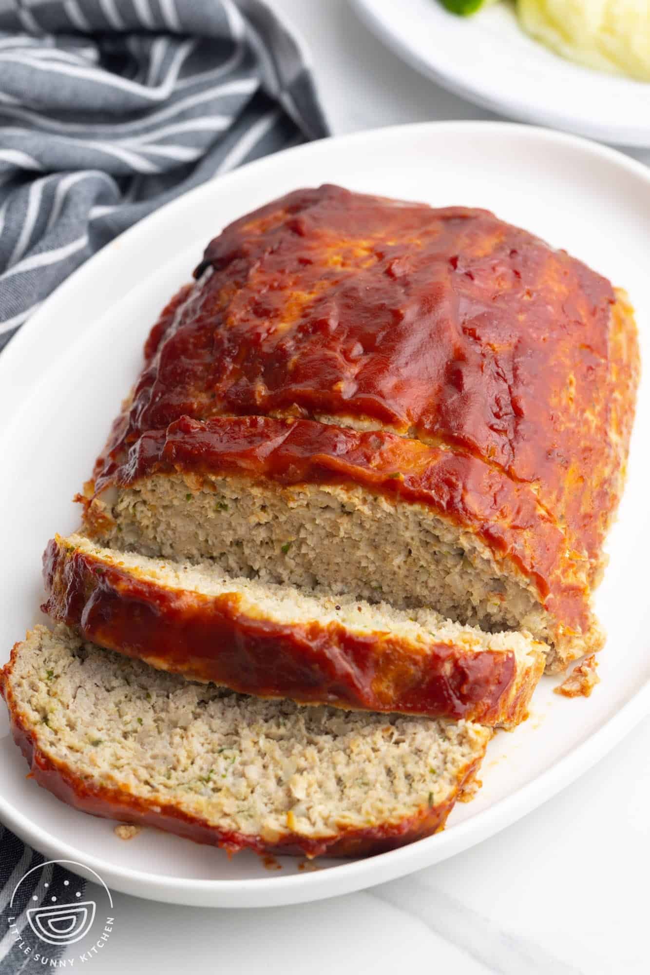 A glazed meatloaf sits on a white platter with two slices cut. The meatloaf appears moist and tender, covered in a shiny ketchup or barbecue sauce. A striped towel is nearby and mashed potatoes are seen in the background.