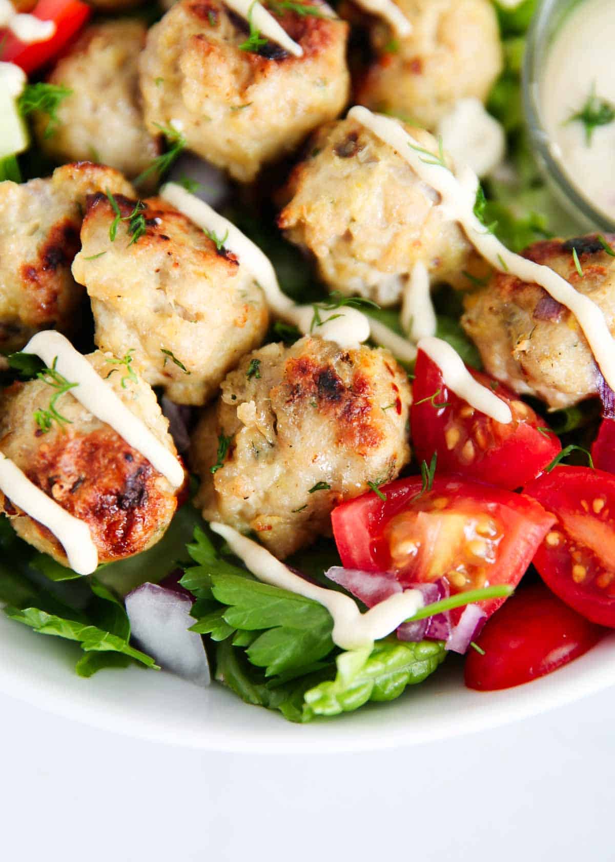 A close-up of a salad bowl with grilled meatballs drizzled with sauce, cherry tomatoes, chopped red onion, greens, and fresh herbs.