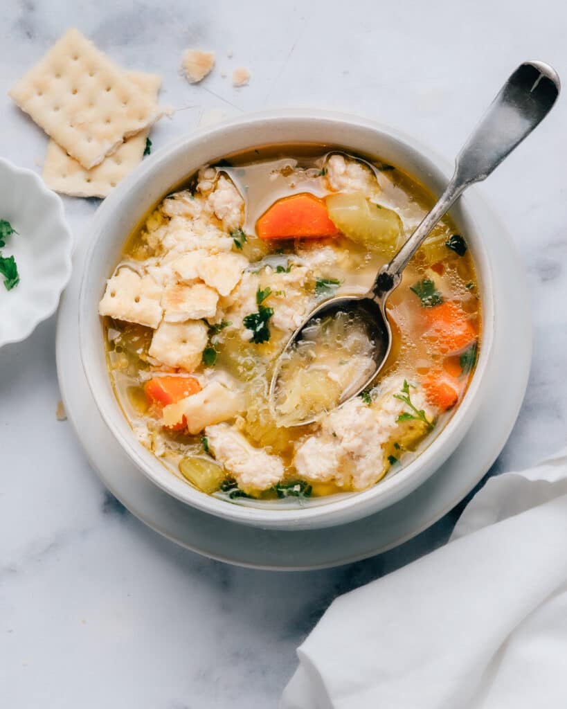 A bowl of chicken soup with carrots, celery, herbs, and crumbled saltine crackers on top. A spoon rests in the bowl, and a plate with crackers is beside it on a white marble surface.